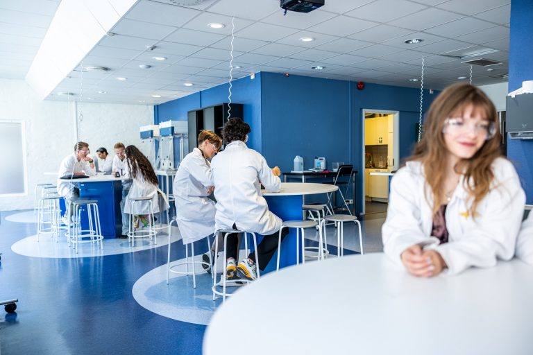  Elever i labbrockar som arbetar i en blå labbsal vid olika bord.