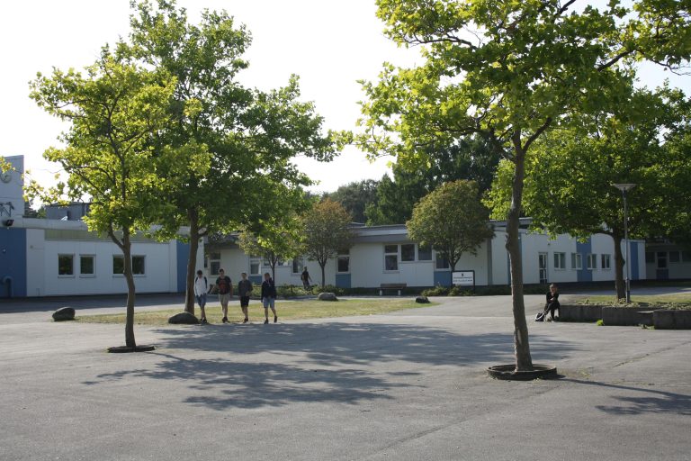 Skolgård med träd, några elever och en vit byggnad i bakgrunden. Huvudingången till Österänggymnasiet.