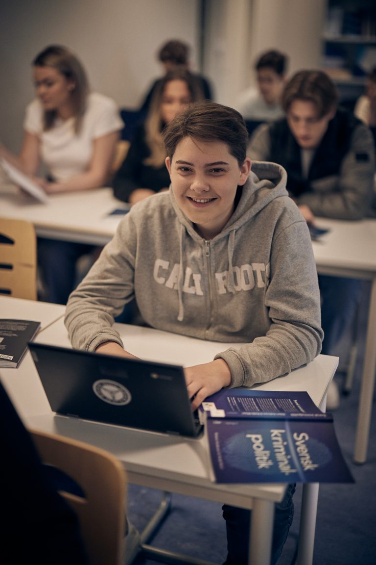Glad elev med en laptop och en bok framför sig i ett klassrum med andra elever i bakgrunden.