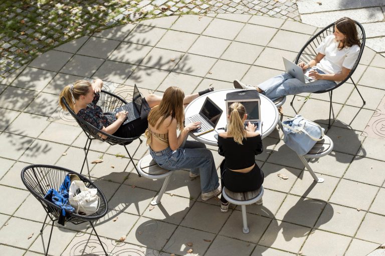 Elever sitter i stolar vid ett bord med laptoppar och samtalar på skolgården.