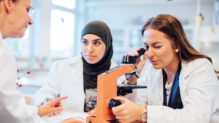 Två elever tittar i ett mikroskop tillsammans med en lärare i en sal. Alla har vita labbrockar. 