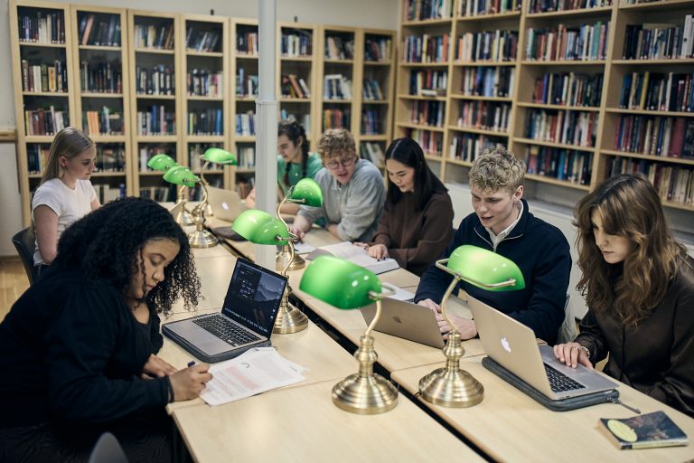 Ett gäng elever sitter koncentrerat och pluggar i skolans bibliotek. Gröna skrivbordslampor lyser upp deras arbeten.