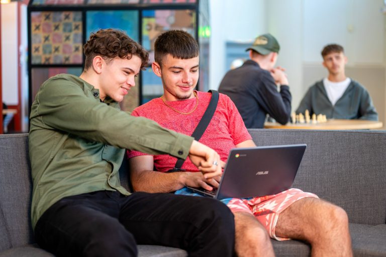 Två glada elever arbetar tillsammans på en laptop. I bakgrunden sitter två andra elever och spelar schack.