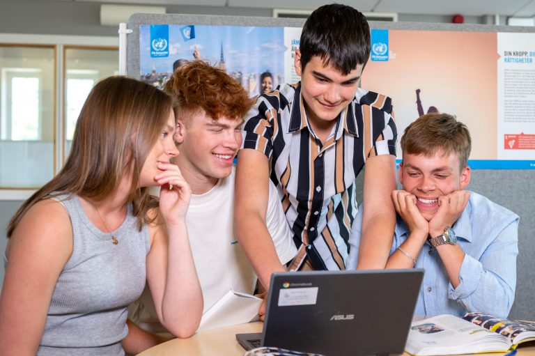 Fyra glada elever löser en uppgift tillsammans på en laptop.