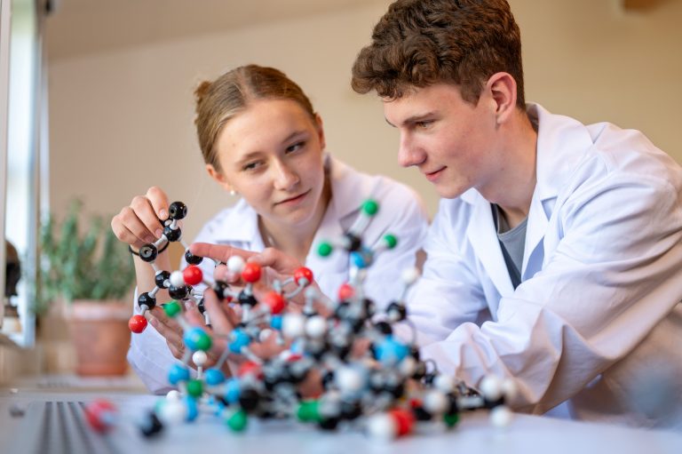 Två koncentrerade elever i labbrockar tittar på en naturvetenskaplig modell. 