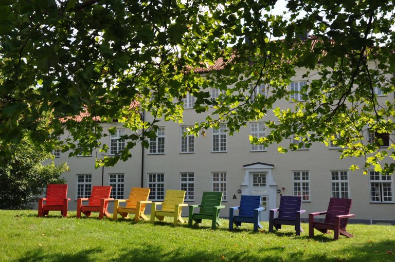 Stolar i regnbågens alla färger på en gräsmatta framför en gul skolbyggnad.