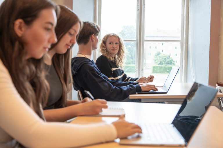 Fyra elever i rad i ett klassrum med bord och laptoppar framför sig. Ett fönster i bakgrunden.