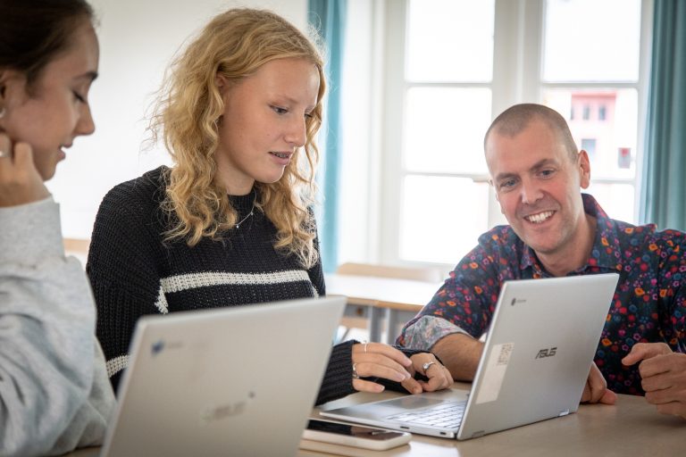 Lärare hjälper två elever som sitter med varsin laptop.