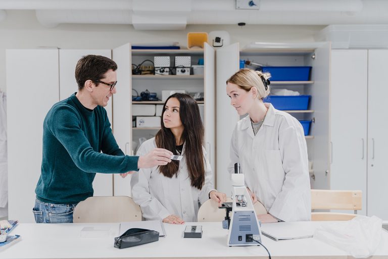 Två elever i labbrockar är i labbet tillsammans med en lärare och tittar i mikroskop