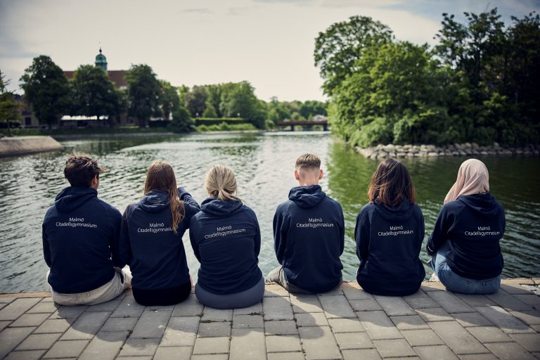 Sex elever sitter på kajkanten med Malmö Citadellsgymnasiums hoodies och tittar ut över kanalen.