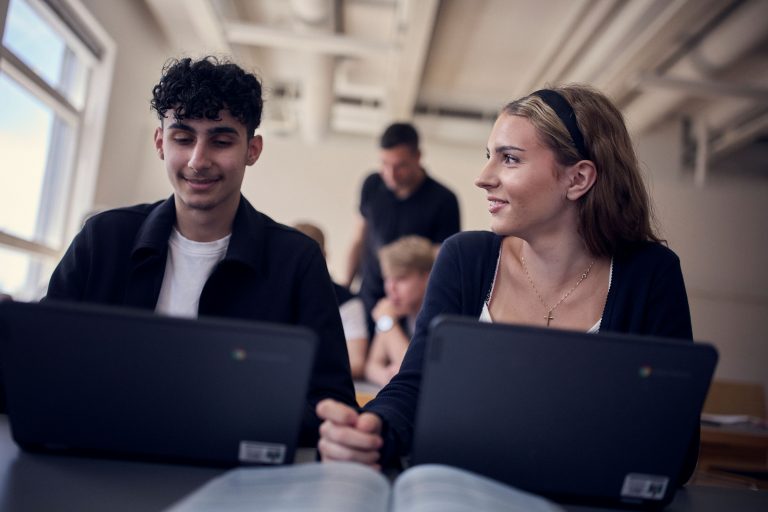 Två glada elever i klassrummet som pluggar med datorer framför sig.