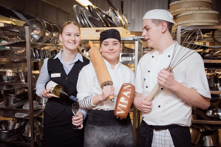 Tre glada elever som visar upp arbetsredskap och uniformer när de serverar, lagar mat och bakar.
