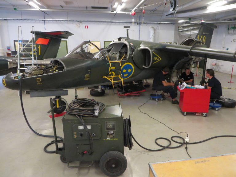 Två elever och en instruktör sitter och diskuterar under vingen på ett av stridsflygplanen i skolans hangar. 