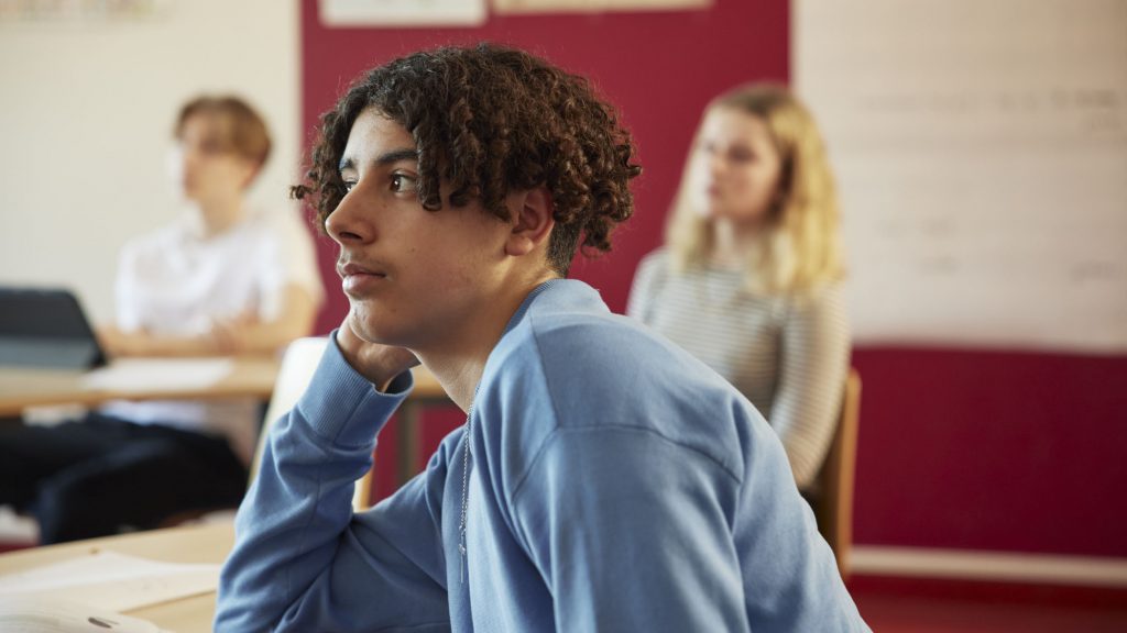 Tonårskille sitter och funderar med huvudet lutat mot ena handen i ett klassrum.
