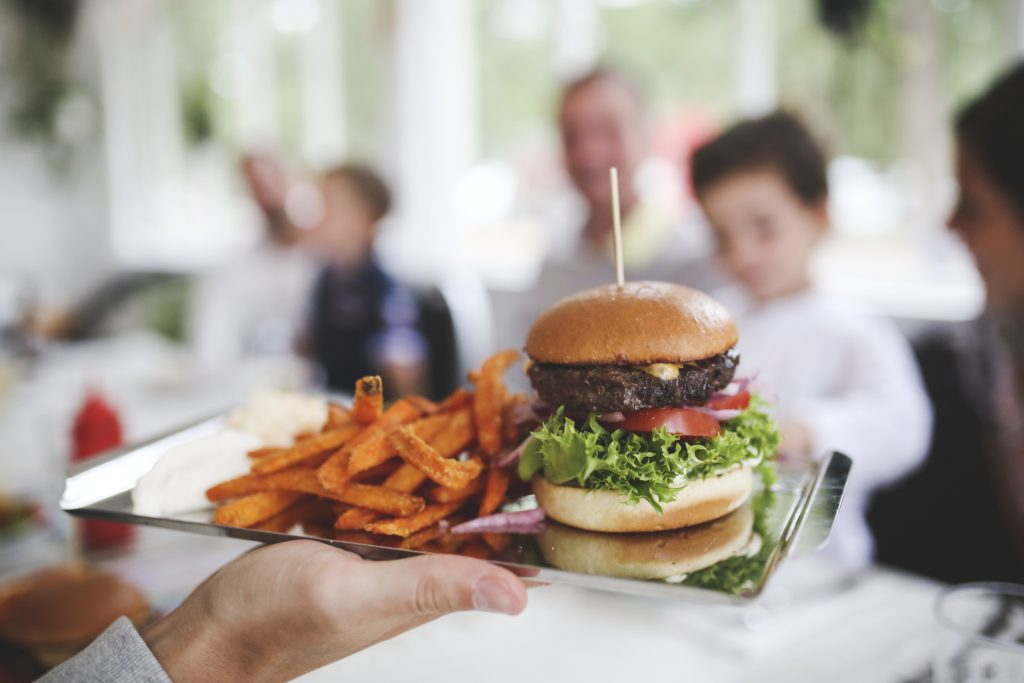 Hand som håller fram ett fat med en hamburgare och sötpotatispommes mot ett sällskap på en restaurang som skymtar suddigt i bakgrunden.