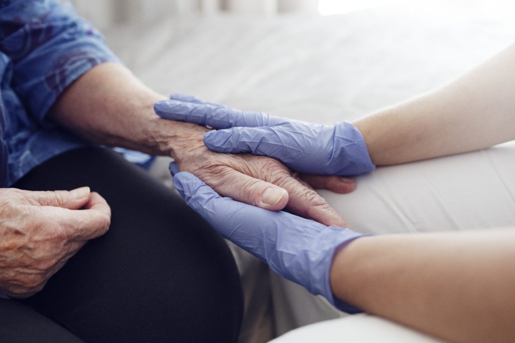 Närbild på händer med plasthandskar på som håller äldre persons hand.