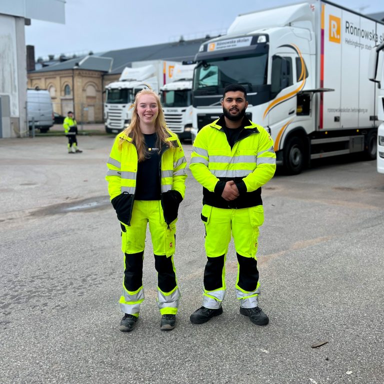 Två elever klädda i yrkeskläder står redo för att köra lastbilar på transportprogrammet.