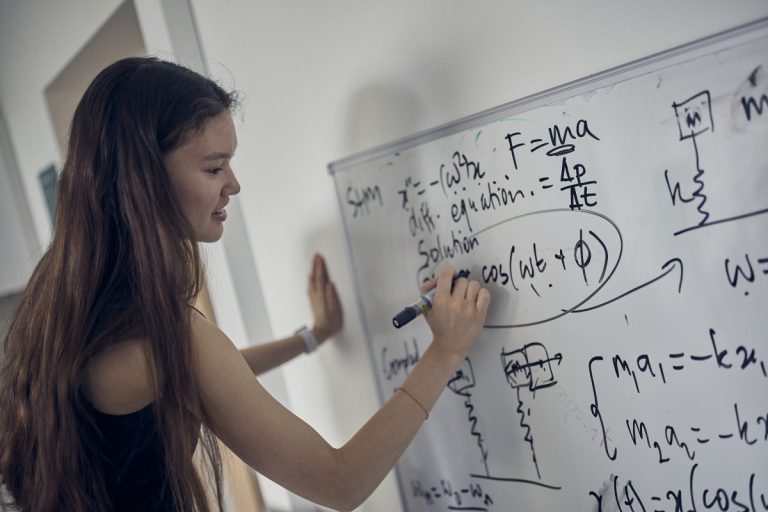 En elev håller på att skriva upp en matematisk formel på en vit tavla i klassrumsmiljö.