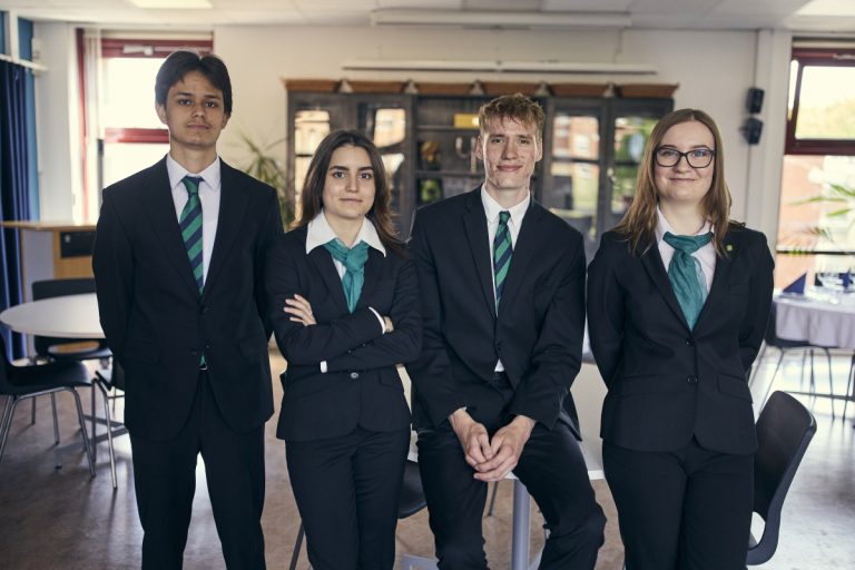 Fyra elever iklädda mörk kavaj och grön slips eller sjal poserar i klassrumsmiljö.