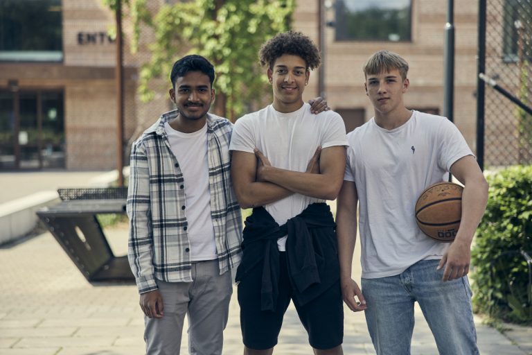 Tre elever poserar tillsammans med en basketboll utomhus i skolgårdsmiljö. 