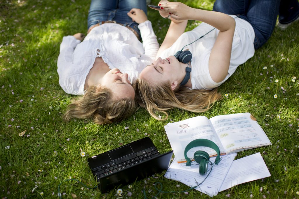 Två glada tonårstjejer ligger på rygg i gräset bredvid dator och skolböcker och läser på en mobiltelefon.